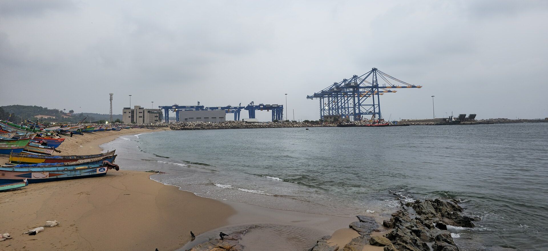 Vizhinjam International Seaport seen across the water from the beach, with Kerala fishing boats drawn up on the sand and the port's gantry cranes on the breakwater beyond.